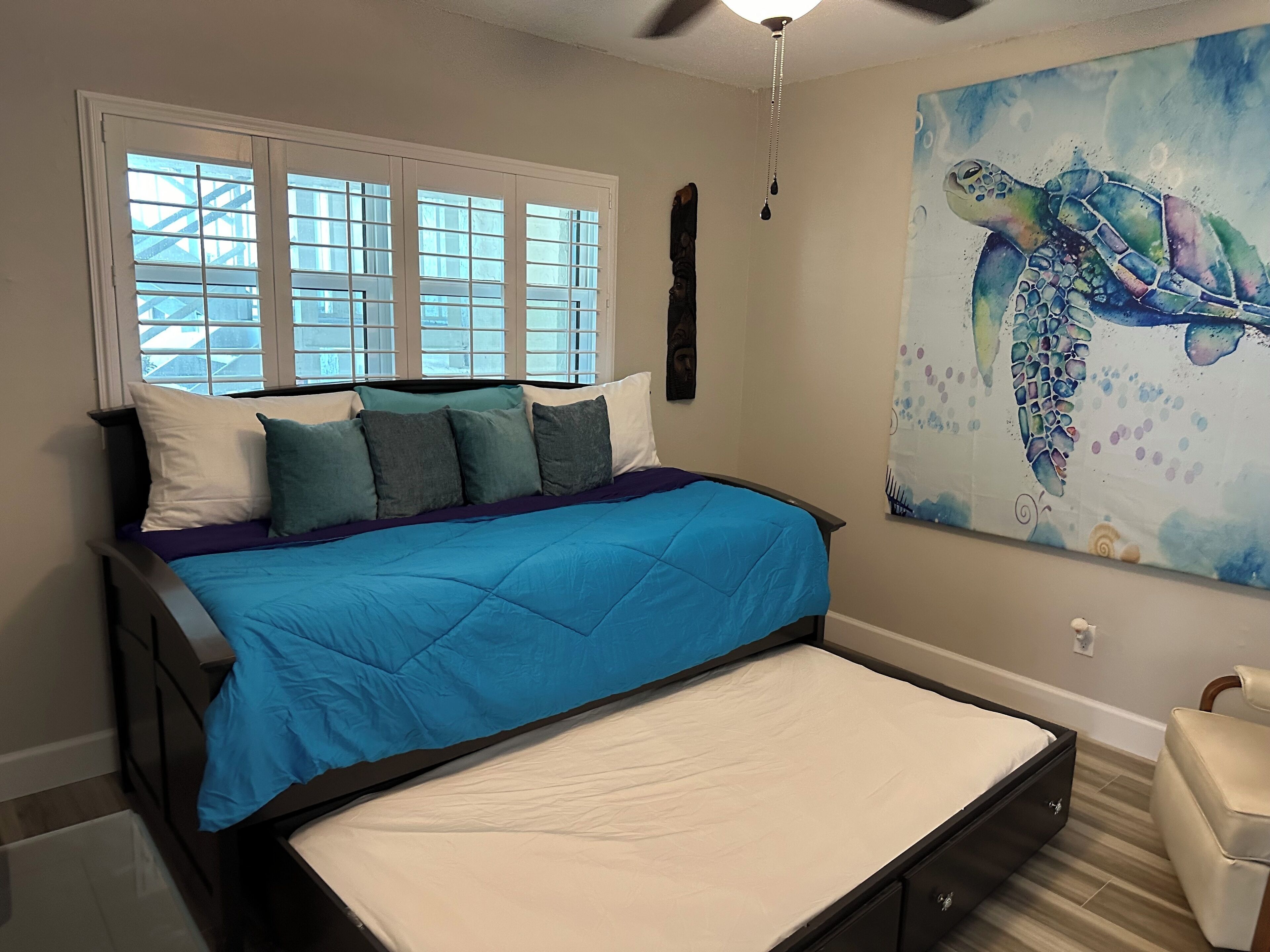Un dormitorio con una cama, ventana, ventilador de techo y un cuadro en la pared.