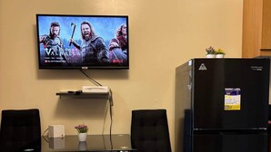 A room with a TV displaying 'Valhalla', a black refrigerator, two black chairs, a glass table, and a clock on the wall. - Apartment hotel in front of biggest mall of asia with resort type amenity pool.  (Pasay)