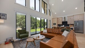 Un espacio habitable amplio con un sofá grande, una mesa de centro y un sillón verde. Se ve una cocina con electrodomésticos modernos al fondo.