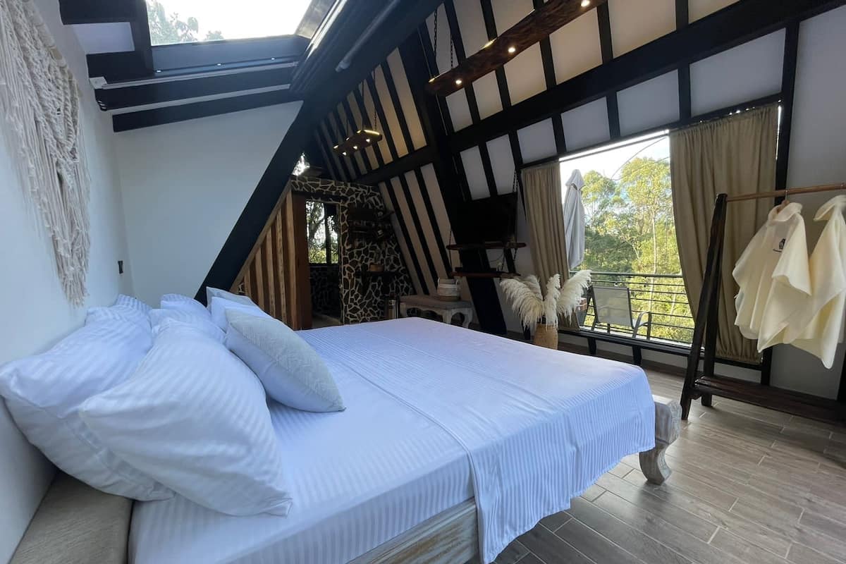 A bedroom with a large bed, wooden beams, a balcony with a view, and a hanging chair.