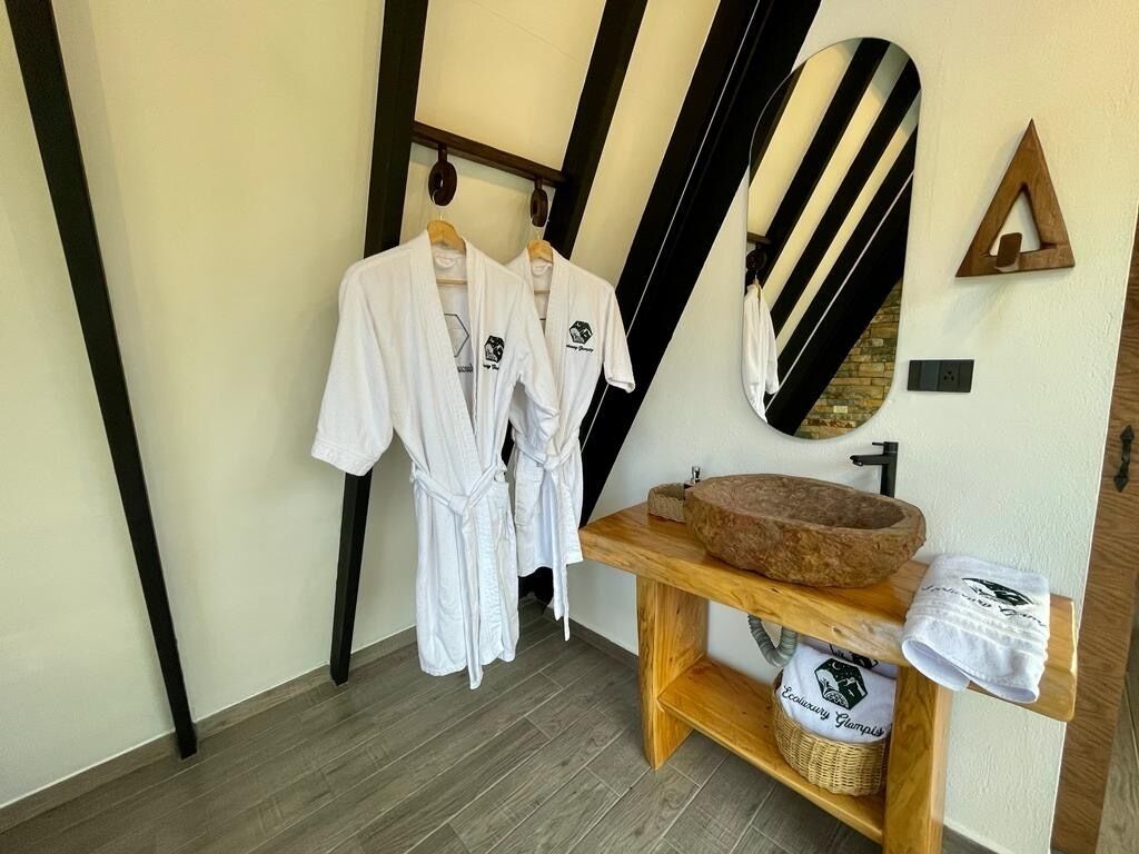 A room with two white robes hanging on a rack, a wooden vanity with a stone basin, and a large mirror with a black and white striped frame.