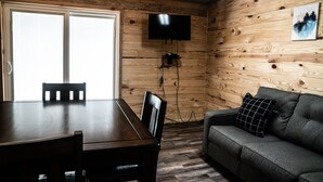 A wooden-paneled room with a TV mounted on the wall, a dining table with chairs, and a sofa with a pillow.