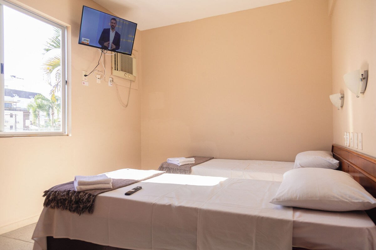 A hotel room with two beds, a TV mounted on the wall, and a window providing a view of palm trees and buildings.