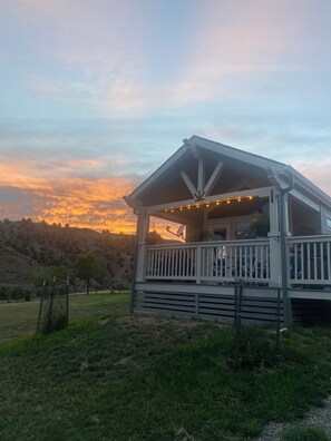 A cabin with a porch, surrounded by a grassy area and trees, under a sunset sky. - Sweet new Tiny House by the Yellowstone River (Gardiner)