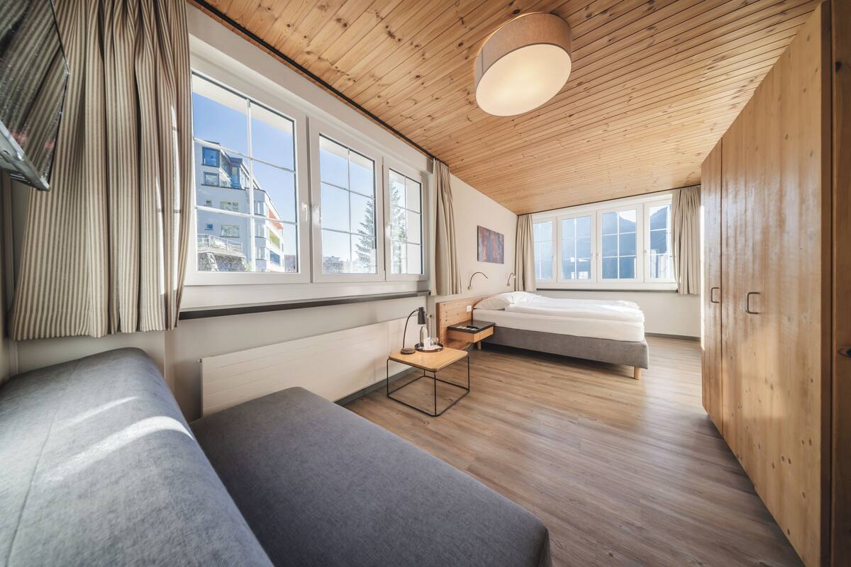 Quarto moderno com teto e pisos de madeira, duas camas, uma escrivaninha e janelas amplas.