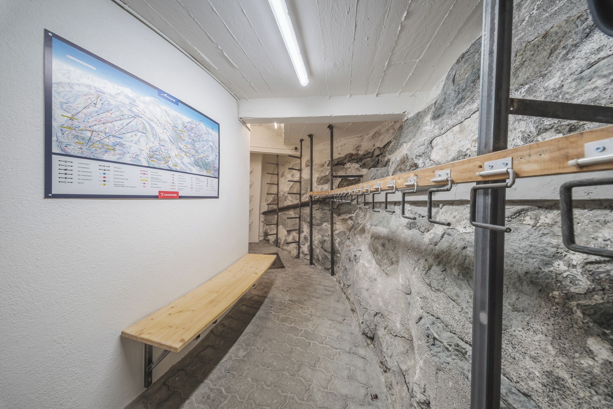 Un couloir avec un banc en bois, une carte accrochée au mur et un présentoir métallique muni de crochets.