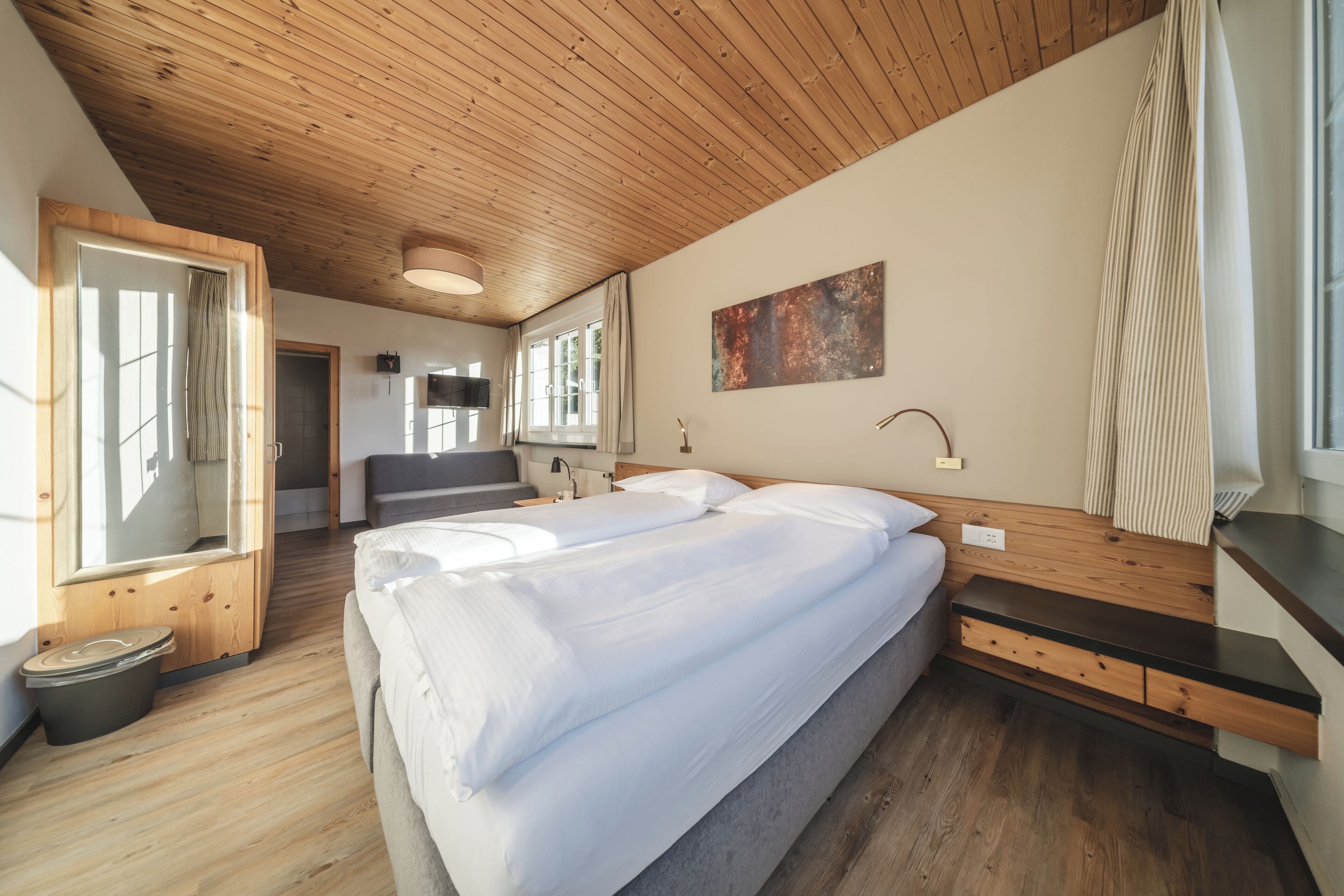 Un dormitorio con techo de madera, una cama con ropa de cama blanca, una mesita de noche, un espejo y una ventana con cortinas.