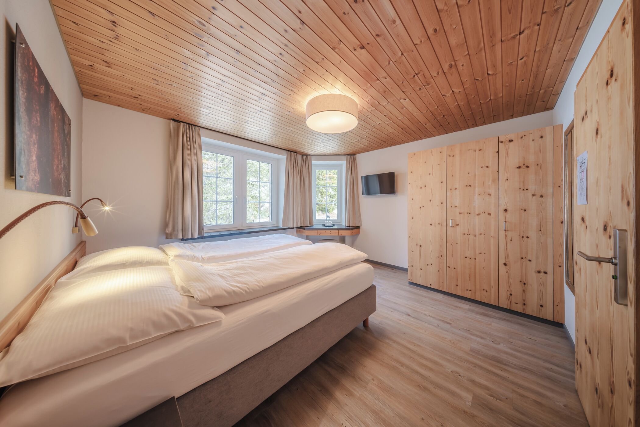 A bedroom with a wooden ceiling, a large bed, a TV mounted on the wall, and a wooden wardrobe.