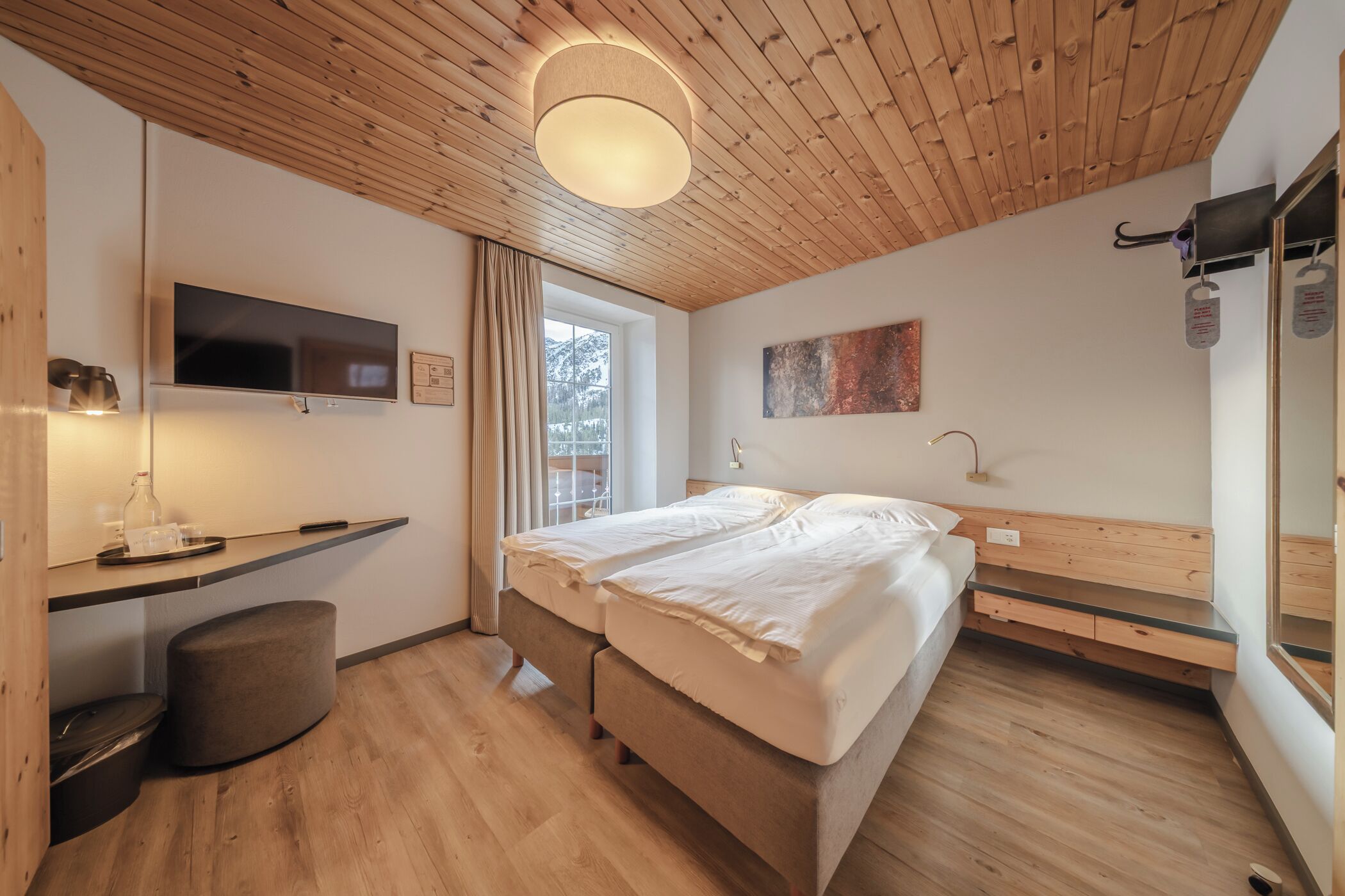Et moderne soveværelse med træloft, seng med hvide sengetøj, et fjernsyn, et skrivebord med en lampe og et vindue med gardiner.