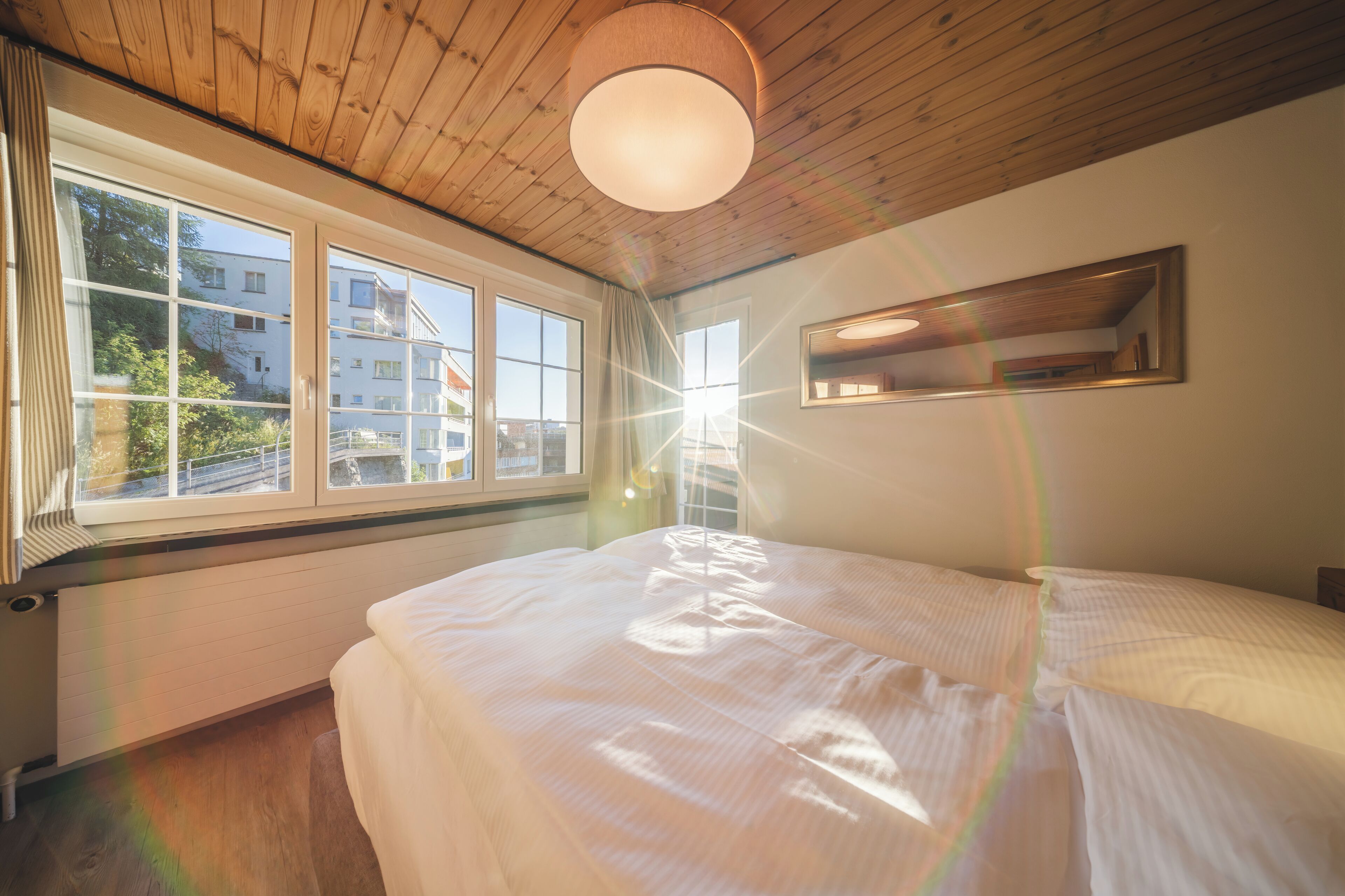 A bedroom with a large window, wooden ceiling, and a bed with white bedding.