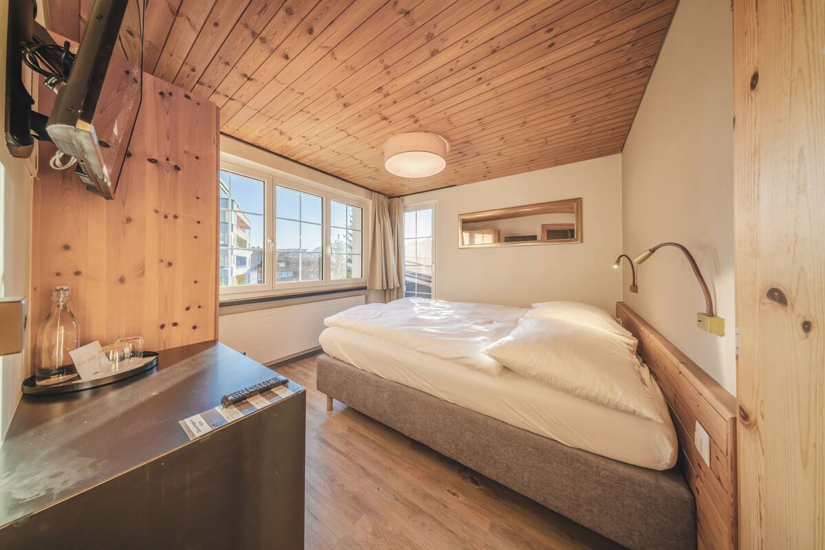 Quarto com teto de madeira, cama com roupa de cama branca, mesa de cabeceira com abajur e janela com cortinas.