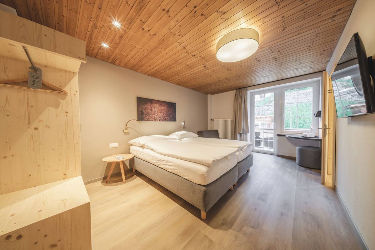 Quarto moderno com teto de madeira, uma cama grande, um banquinho pequeno e uma janela com vista para o exterior.