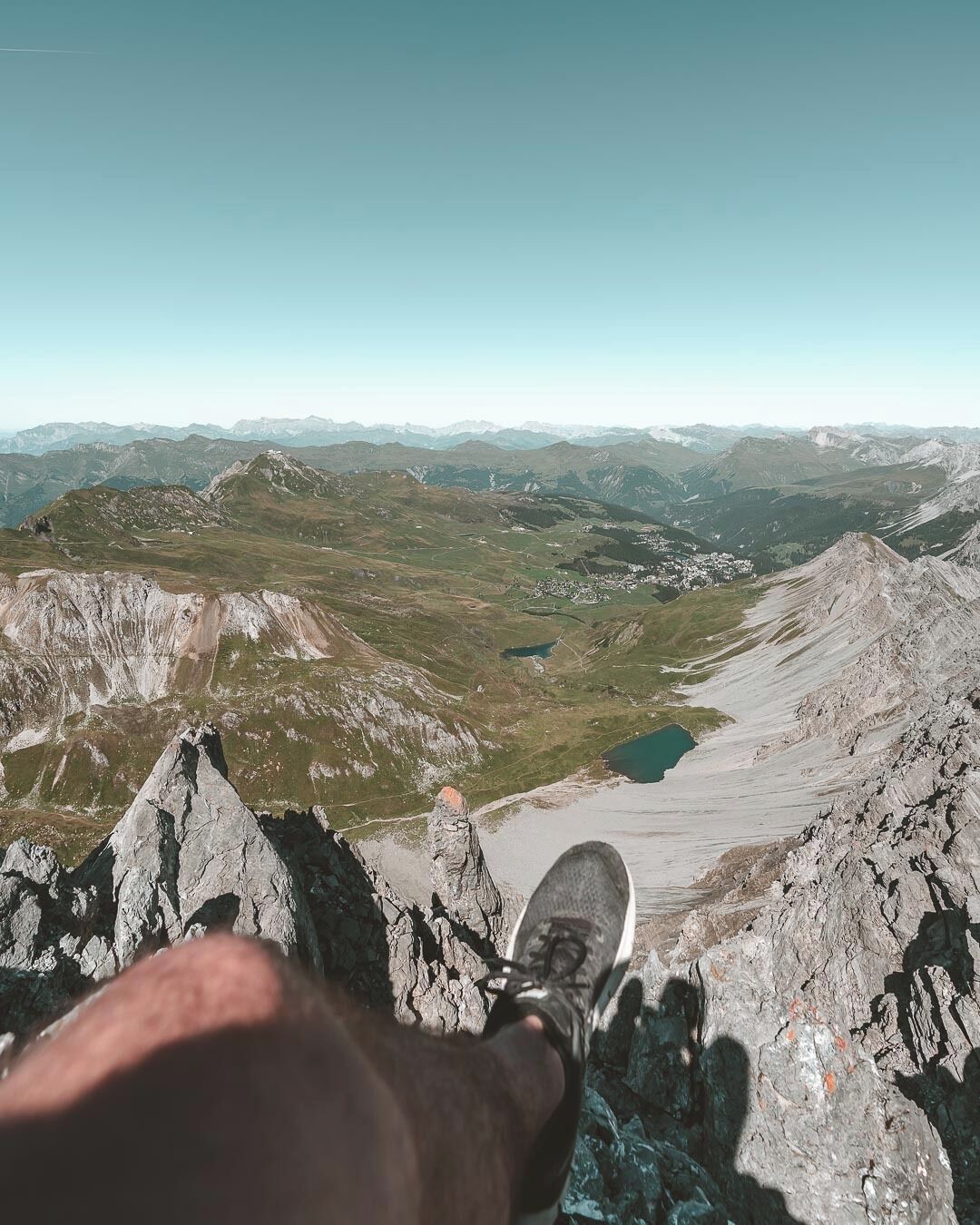 A person's foot on a mountain peak overlooking a valley with a lake and distant mountains.
