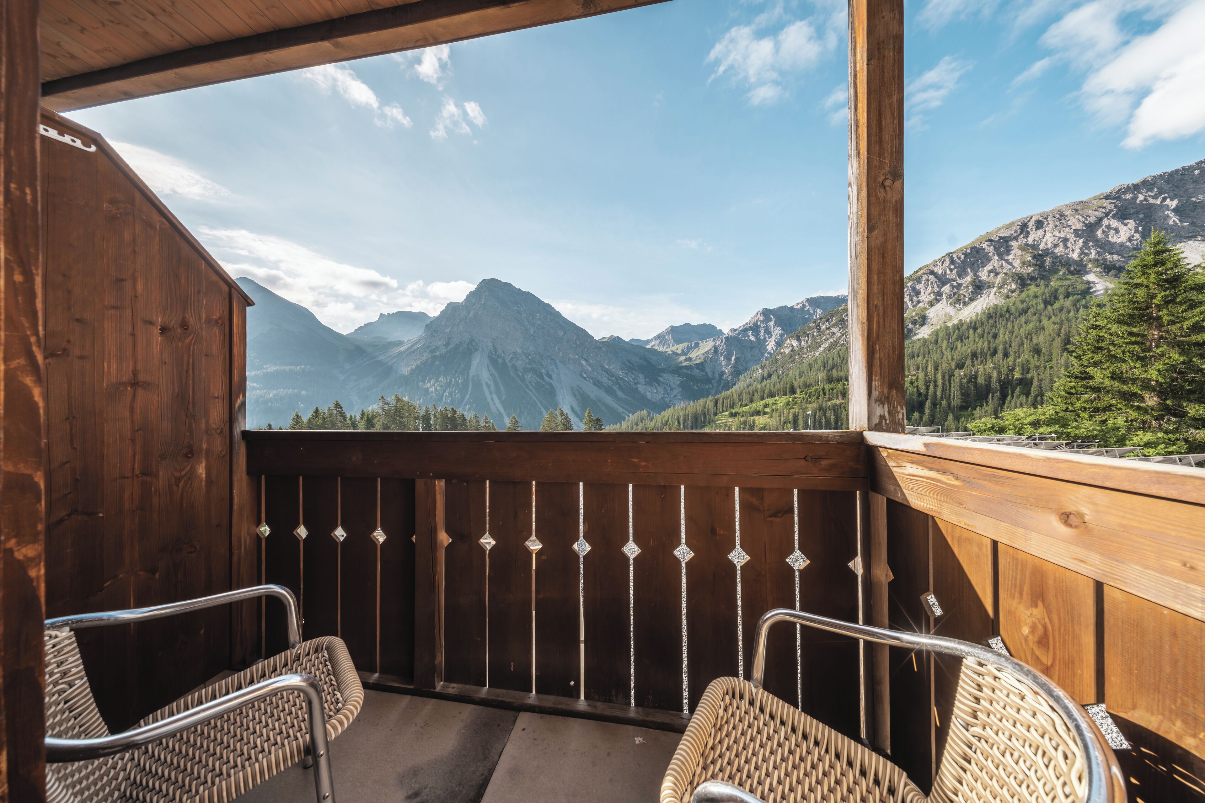 Un balcon en bois agrémenté de chaises en osier, offrant une vue sur les montagnes et la forêt.