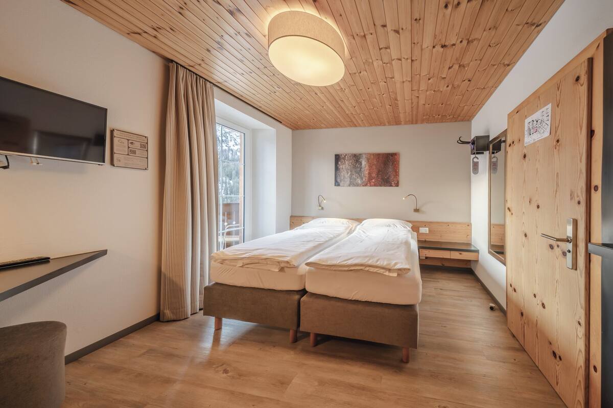 Quarto moderno com teto de madeira, cama com roupa de cama branca, televisão fixada na parede e janela com cortinas.