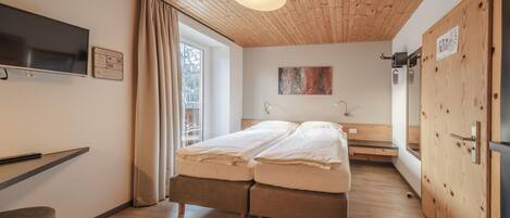 Un dormitorio moderno con techo de madera, cama con ropa blanca, televisor montado en la pared y ventana con cortinas.