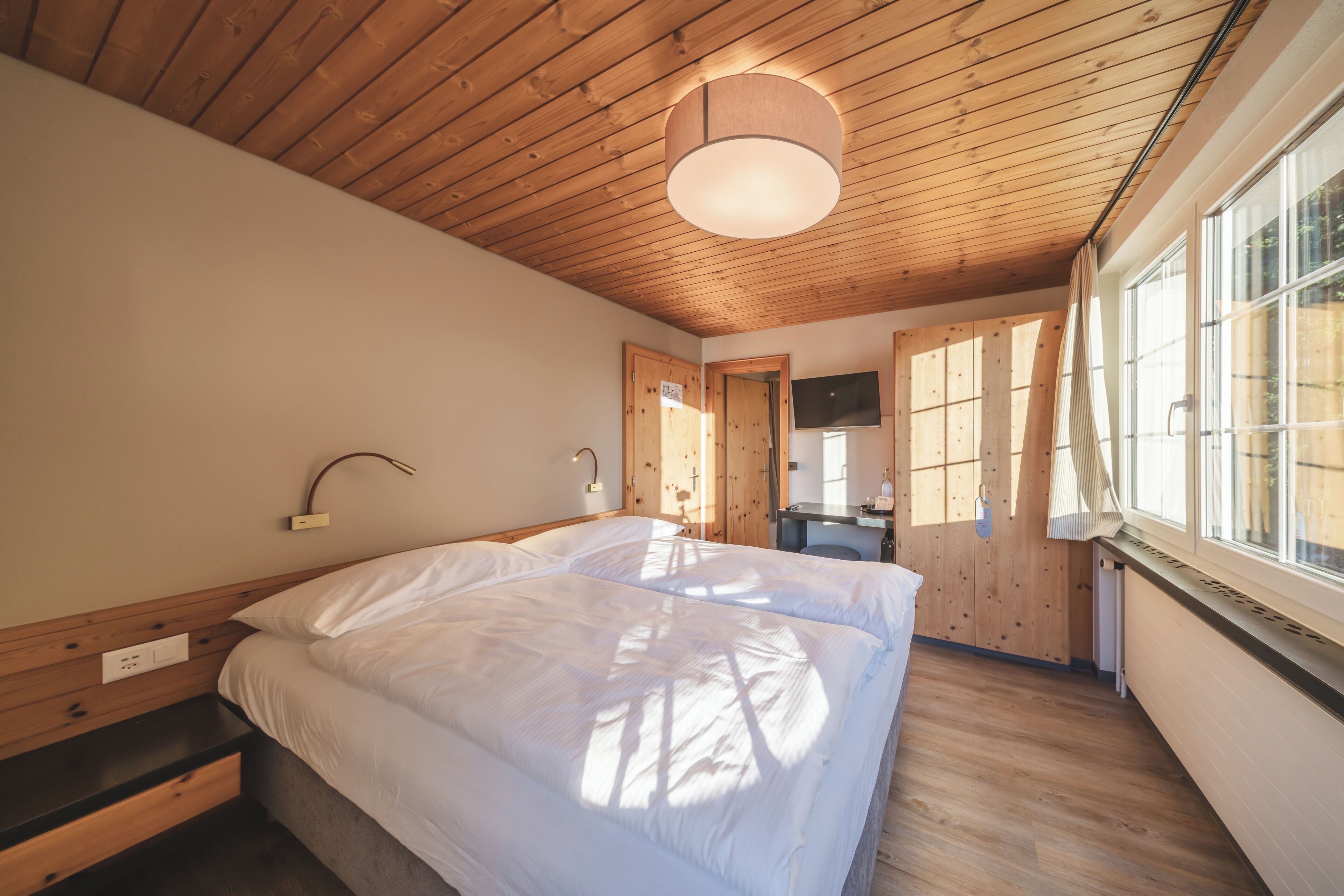 A bedroom with a wooden ceiling, a bed with white bedding, a bedside table, a desk, and a window with a view.