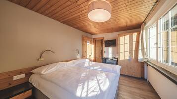 A bedroom with a wooden ceiling, a bed with white bedding, a bedside table, a desk, and a window with a view.