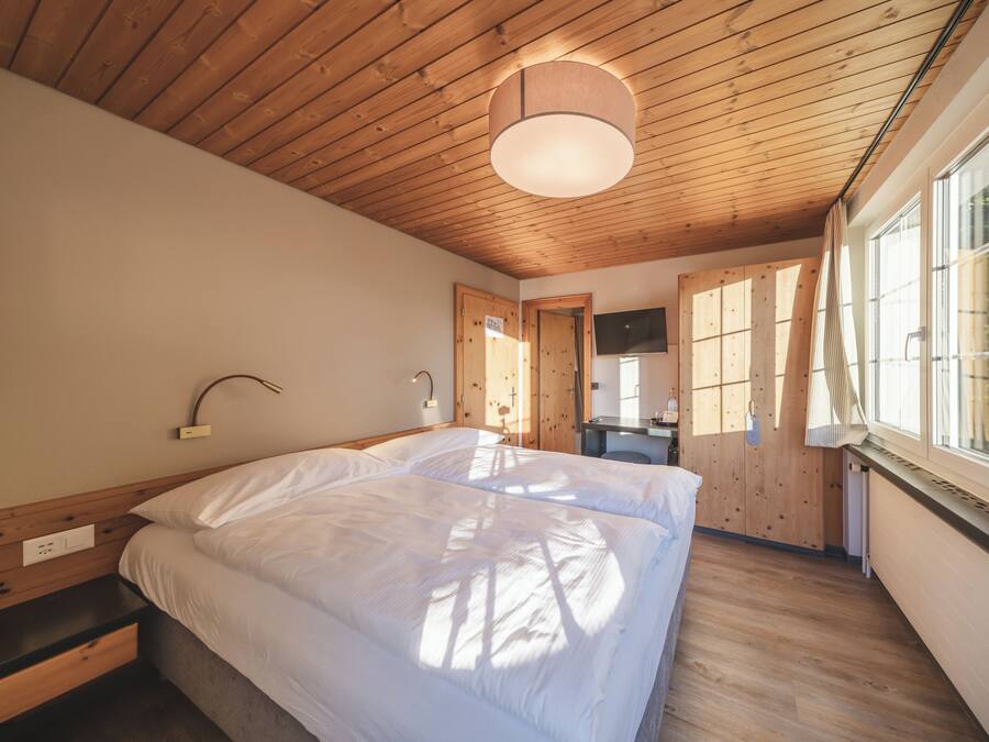A bedroom with a wooden ceiling, a bed with white bedding, a bedside table, a desk, and a window with a view.