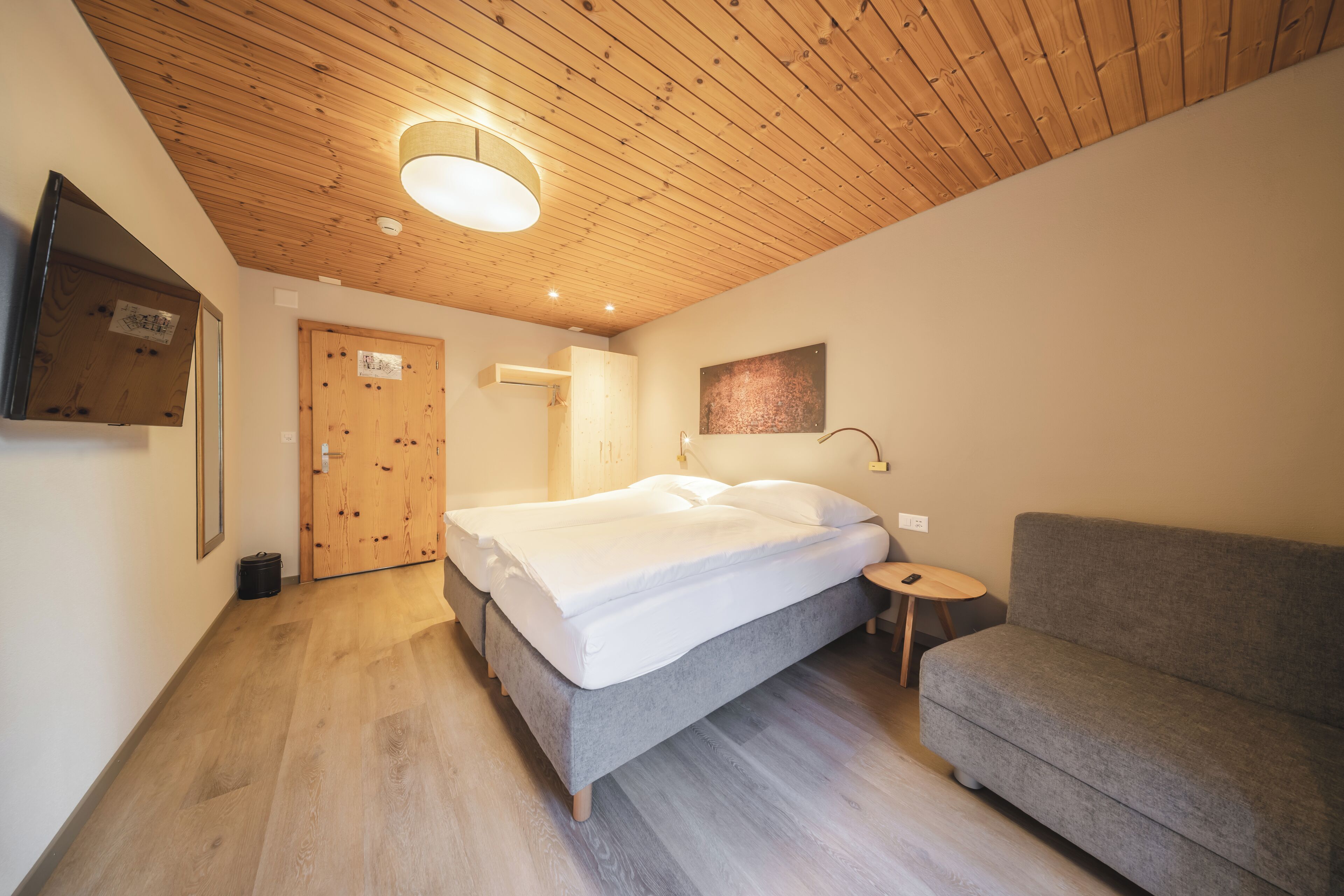 A bedroom with a wooden ceiling, a bed, a grey sofa, and a small table.