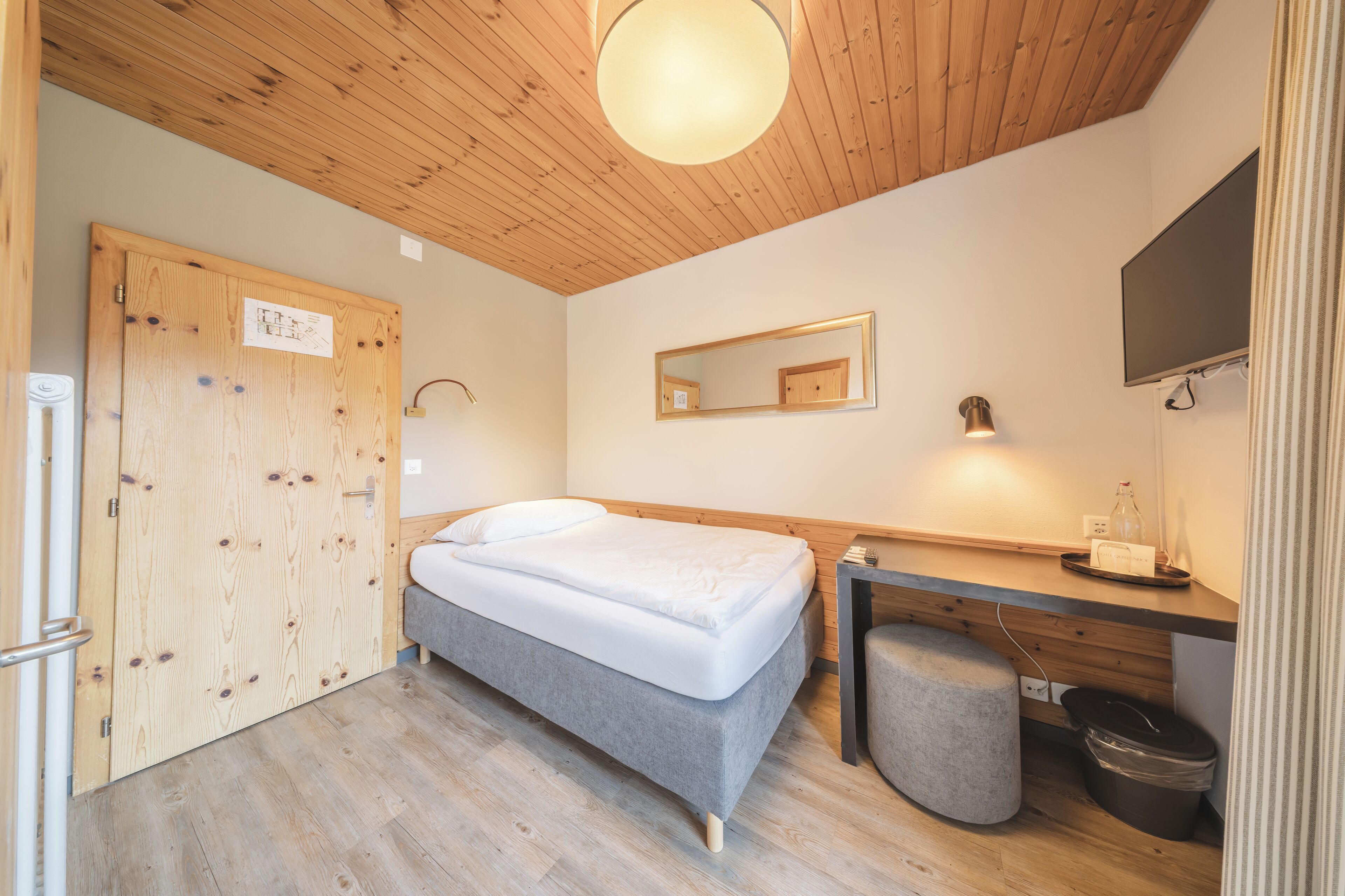 A bedroom with a bed, wooden ceiling, a desk with a chair, a mirror, and a television.