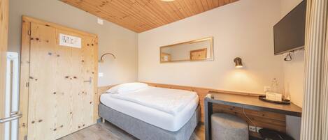 A bedroom with a bed, wooden ceiling, a desk with a chair, a mirror, and a television.
