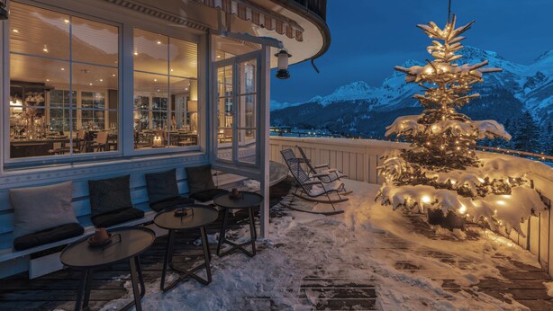 Un área de estar al aire libre cubierta de nieve con mesas y sillas, un árbol de Navidad y vistas a montañas nevadas.