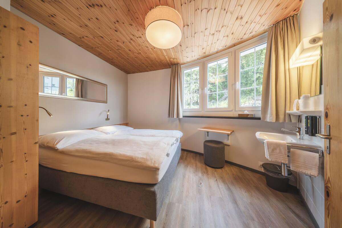 Quarto com teto de madeira, uma cama grande, mesa de cabeceira, espelho, janela com cortinas e banheiro com pia e porta-toalhas.