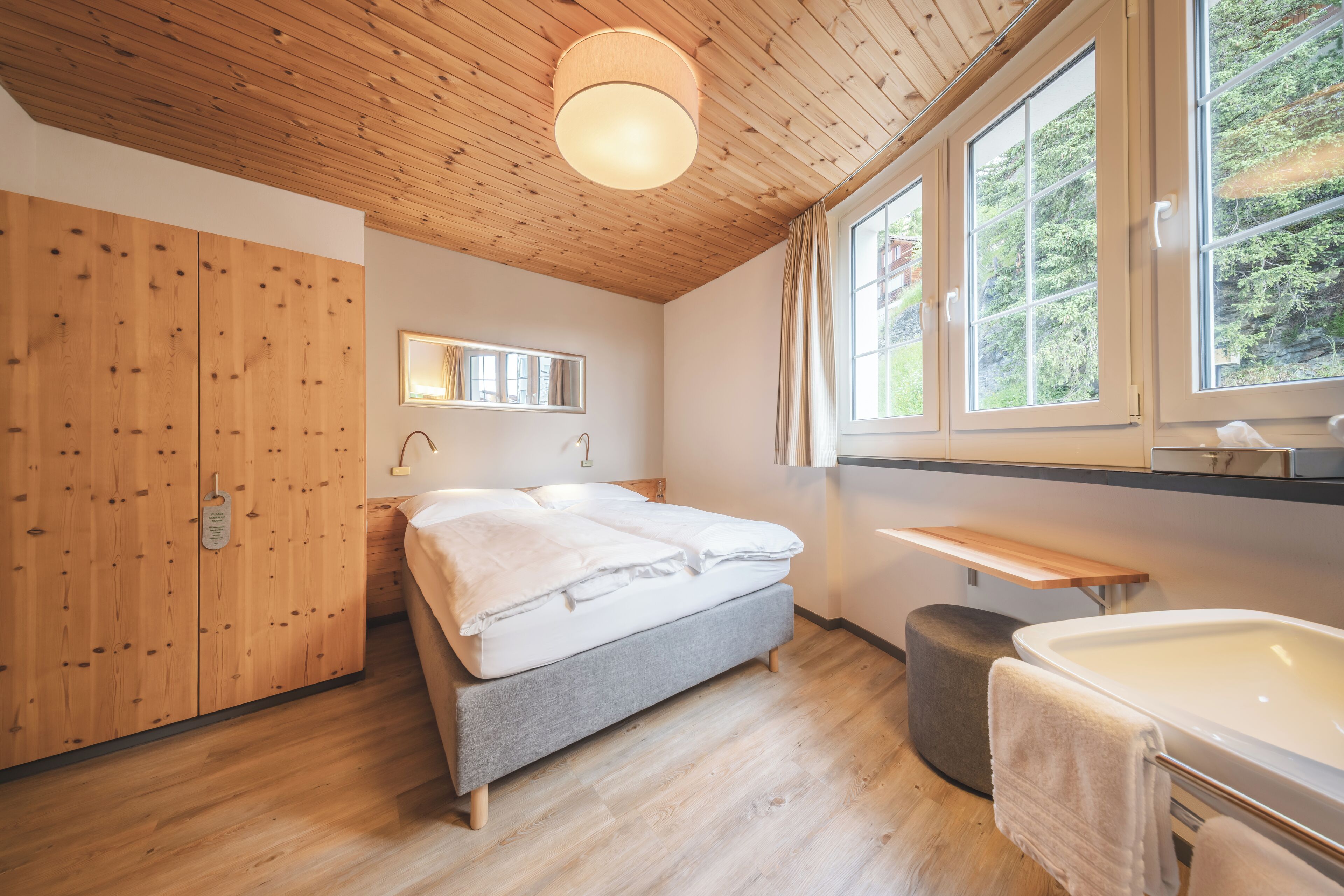 A bedroom with a wooden ceiling, a bed, a bedside table, a window, and a bathroom with a bathtub.