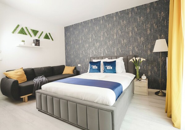 Une chambre à coucher moderne avec un lit gris, un canapé noir, une petite table de chevet avec une lampe et une étagère murale agrémentée d’objets décoratifs.