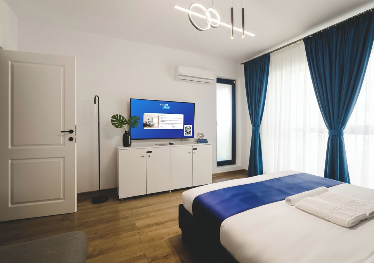 A modern hotel room with a bed, a television, a plant, and a window with curtains.