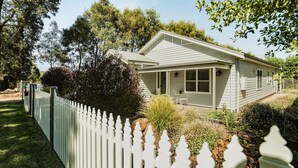 A single-story house with a covered porch, surrounded by a white picket fence and lush greenery. - Cow Bell Cottage, quaint country retreat in Trentham (Trentham)