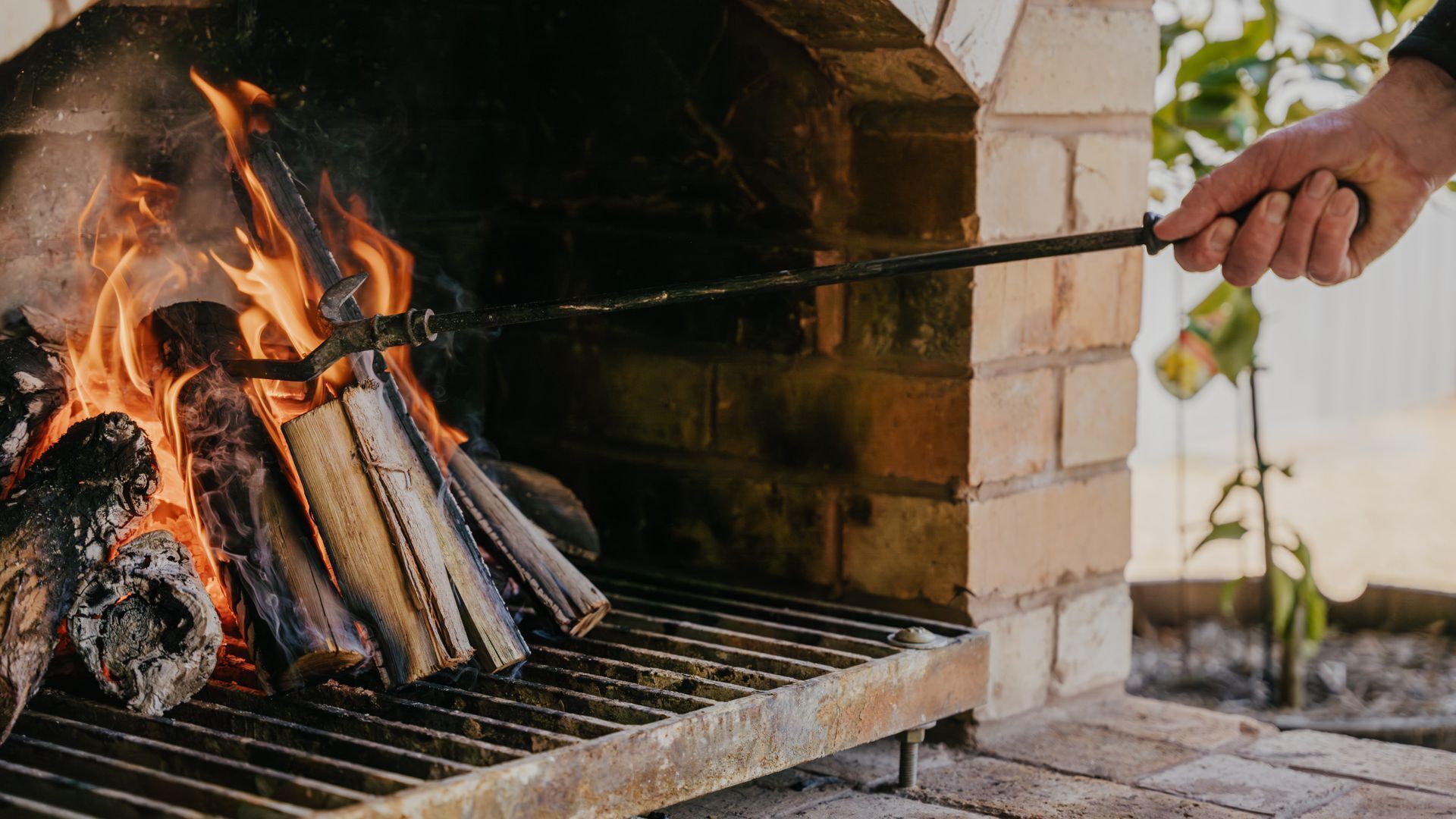 A person using a long-handled tool to manage wood and flames in a brick-built outdoor grill.