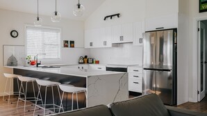 A modern kitchen with white cabinets, a stainless steel refrigerator, and a marble island with bar stools. - Cow Bell Cottage, quaint country retreat in Trentham (Trentham)