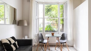 Un salón acogedor con un sofá gris oscuro, una mesita con dos sillas, una lámpara y vistas a zonas verdes a través de amplios ventanales.