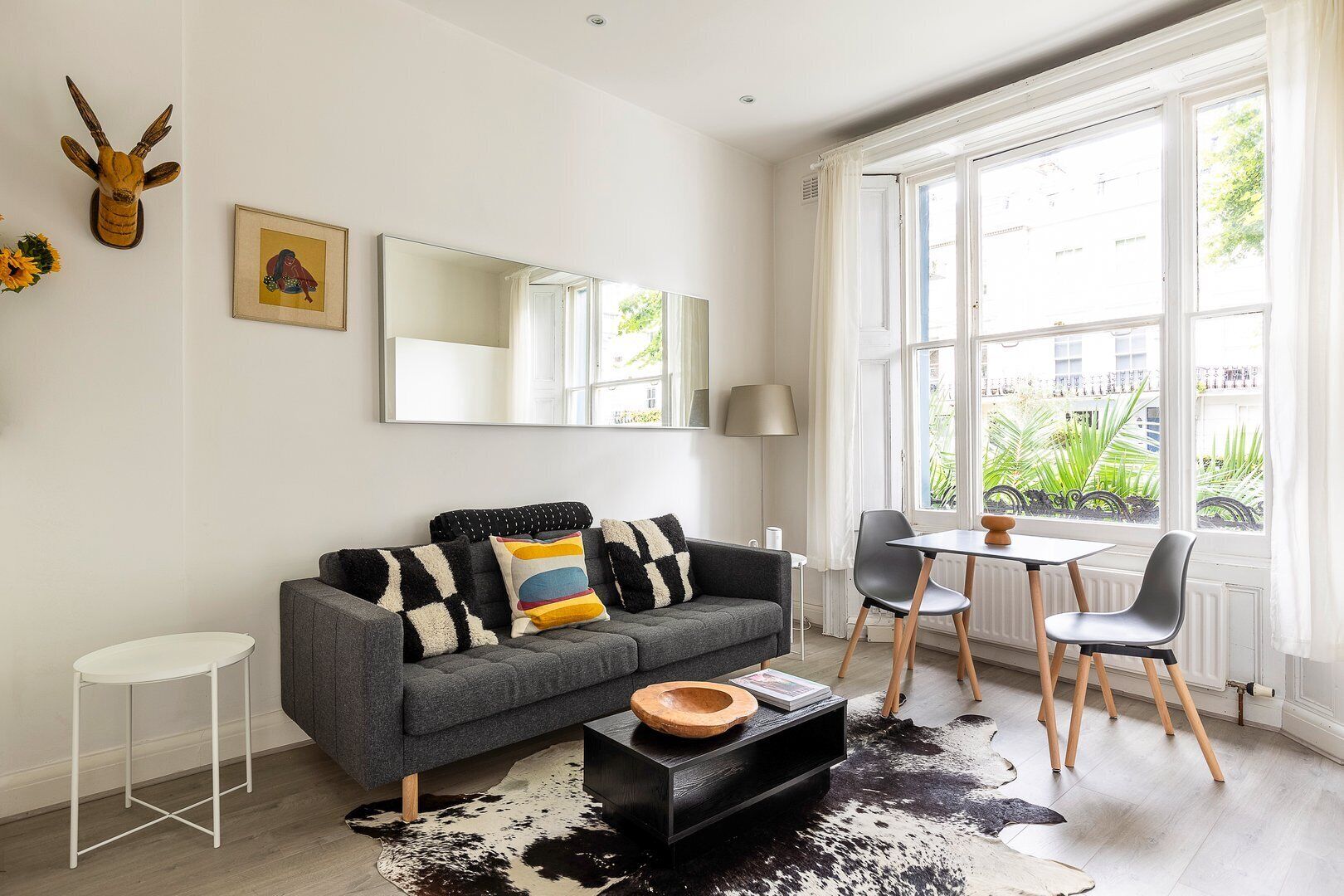 A modern living room with a grey sofa, black and white patterned cushions, a black coffee table, and a cowhide rug. There is a dining table with chairs, a lamp, and a large window with a view of greenery outside.