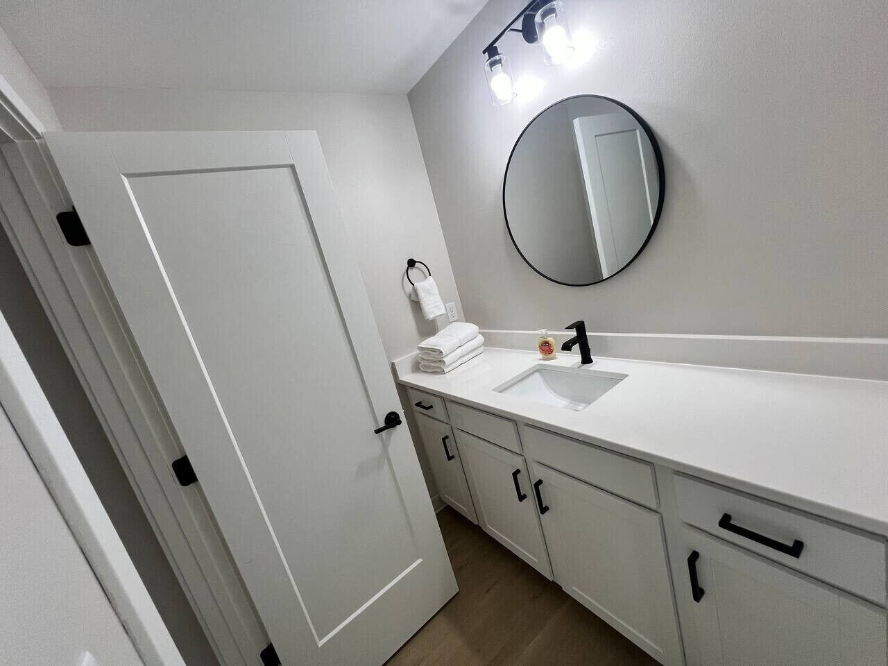 Une salle de bain avec un meuble-lavabo blanc, un miroir rond et un évier. On y trouve deux armoires avec des poignées noires et un porte-serviettes avec une serviette blanche.