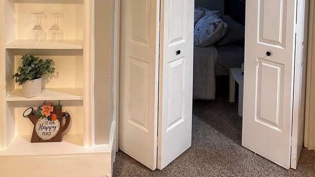 A room with a white door, a shelf with decorative items, and a bed with a headboard. - Modern Tiny Home Escape (Garfield Heights)
