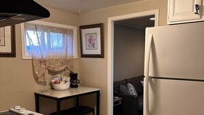 A compact kitchen with a refrigerator, a stove, a small table with a bowl and a coffee maker, and a window with a decorative curtain. - Modern Tiny Home Escape (Garfield Heights)