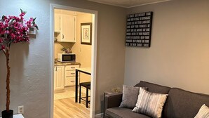 A living room with a brown sofa, a small table, and a potted plant. There is a kitchen visible through an open doorway. - Modern Tiny Home Escape (Garfield Heights)