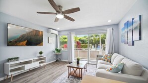A modern living room with a ceiling fan, a large flat-screen TV, a white sofa, and a view of the outdoors through large windows. - NEW! FULLY UPDATED ground floor condo with AC! (Kapaʻa)