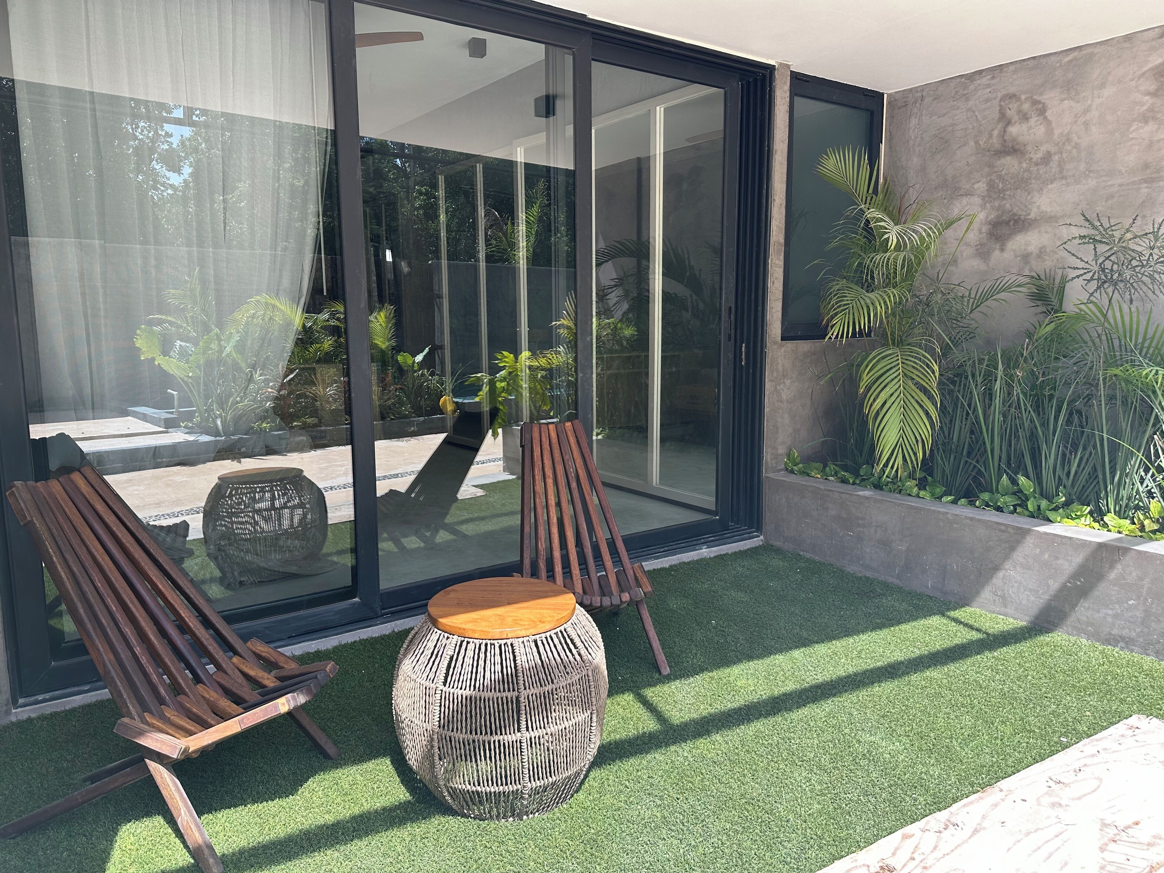 A modern outdoor seating area with two wooden chairs and a woven stool, surrounded by a glass door leading to a garden with various plants.