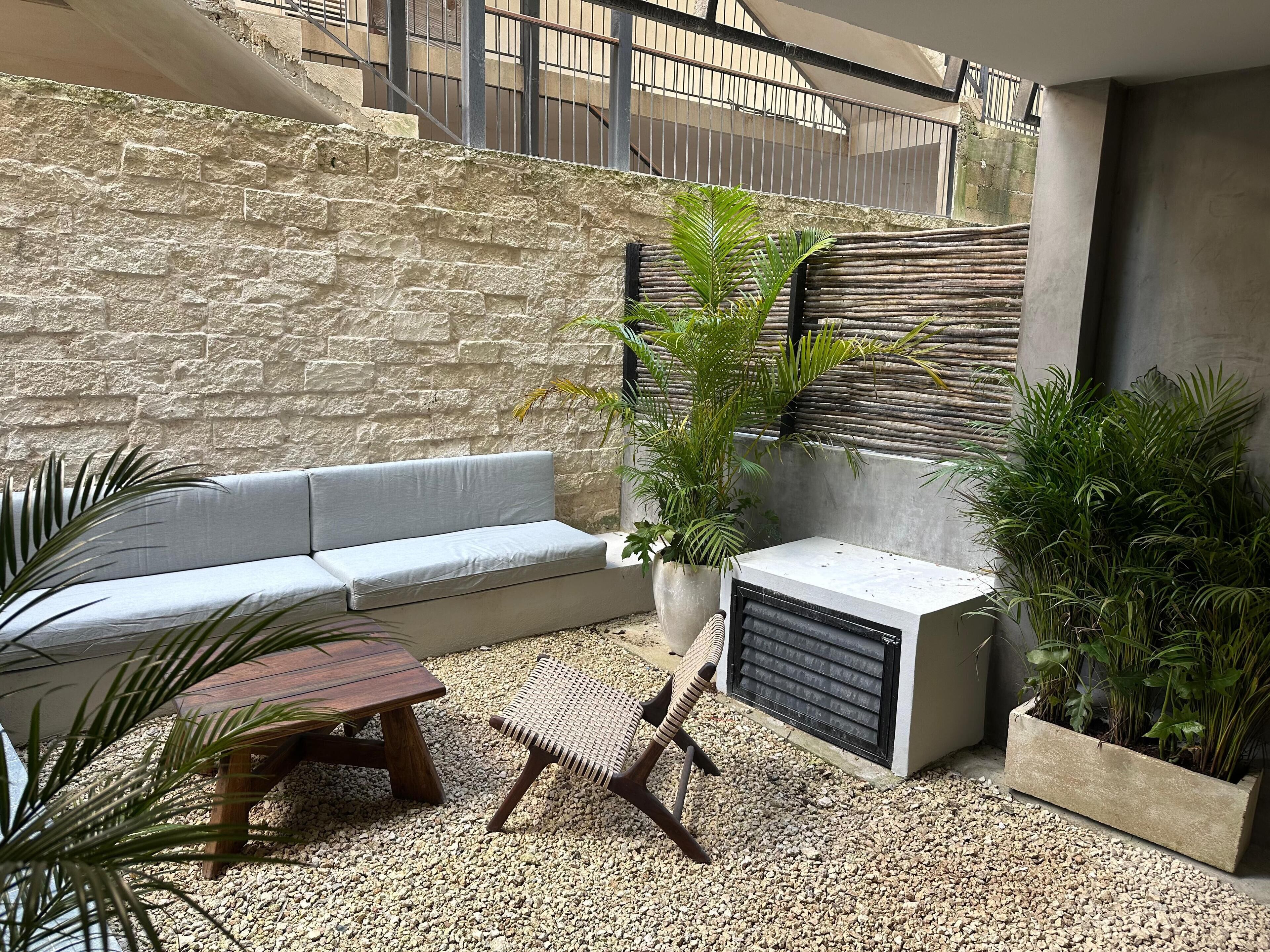 Een patio met een stenen muur, een grijze bank, een houten tafel, een geweven stoel en potplanten.