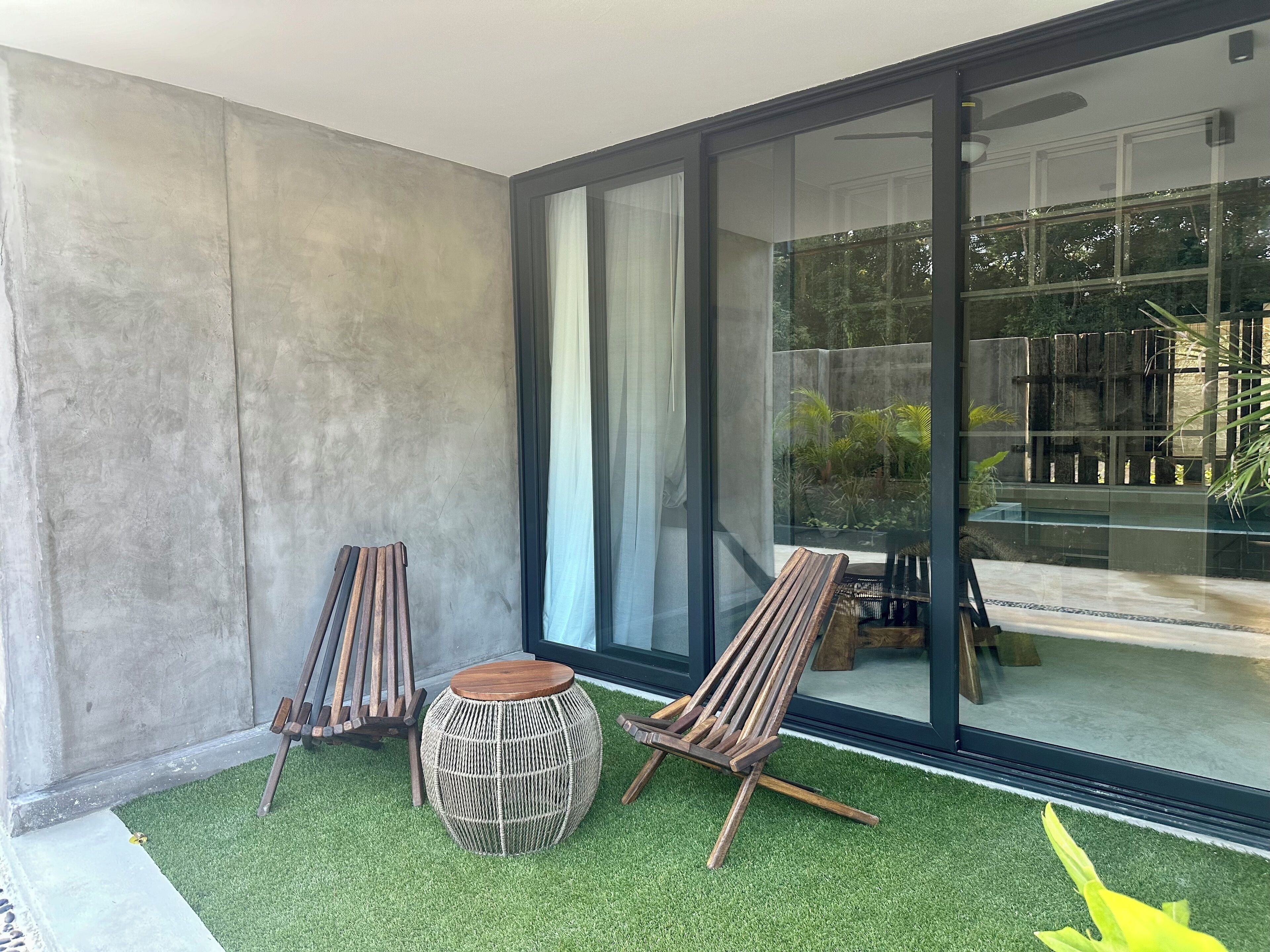 Un espace extérieur moderne aménagé avec deux chaises en bois et un tabouret en osier, le tout posé sur une pelouse artificielle, à côté d’une porte vitrée donnant sur un balcon avec vue sur la verdure.