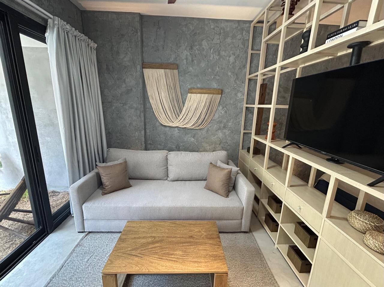 A modern living room with a grey sofa, a wooden coffee table, a TV on a white stand, and a hammock on the wall.
