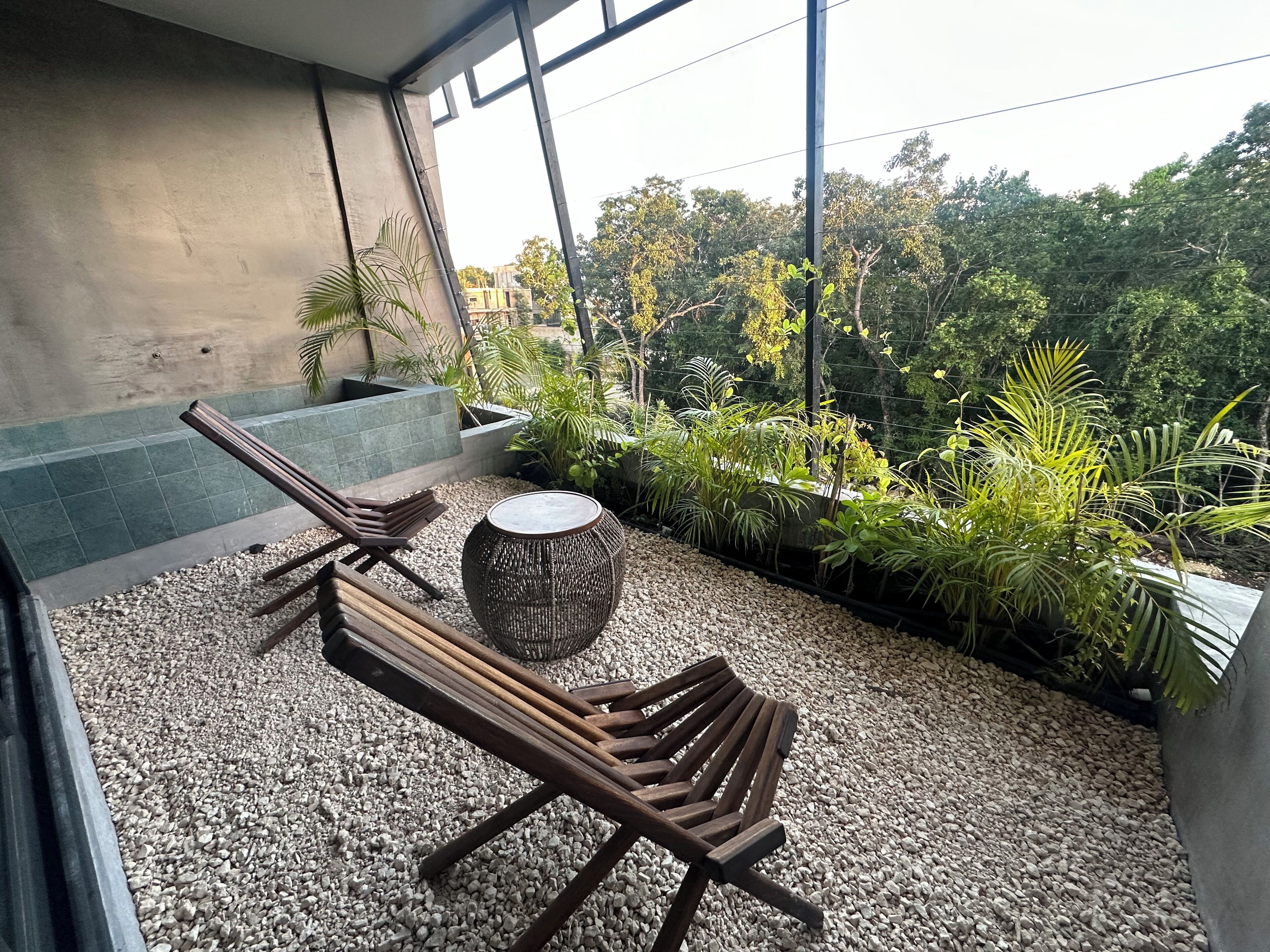 A balcony with two wooden lounge chairs, a woven stool, and a small table. The balcony has a gravel floor and is surrounded by lush greenery and a view of trees.