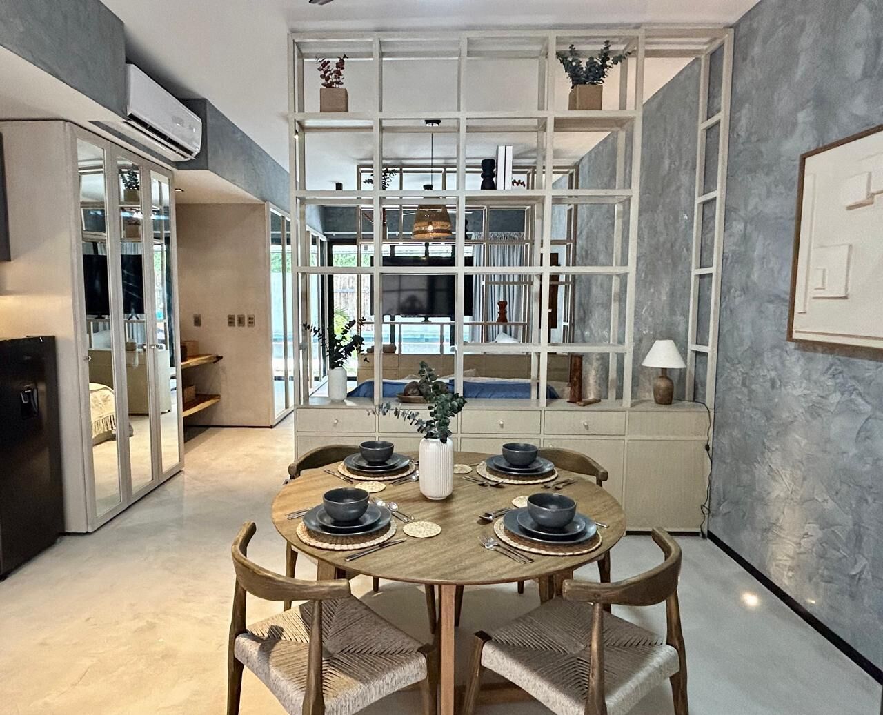 Un coin repas avec une table ronde en bois, dressée pour quatre personnes, entourée de chaises en osier. Un vase blanc contenant une plante est posé sur la table.