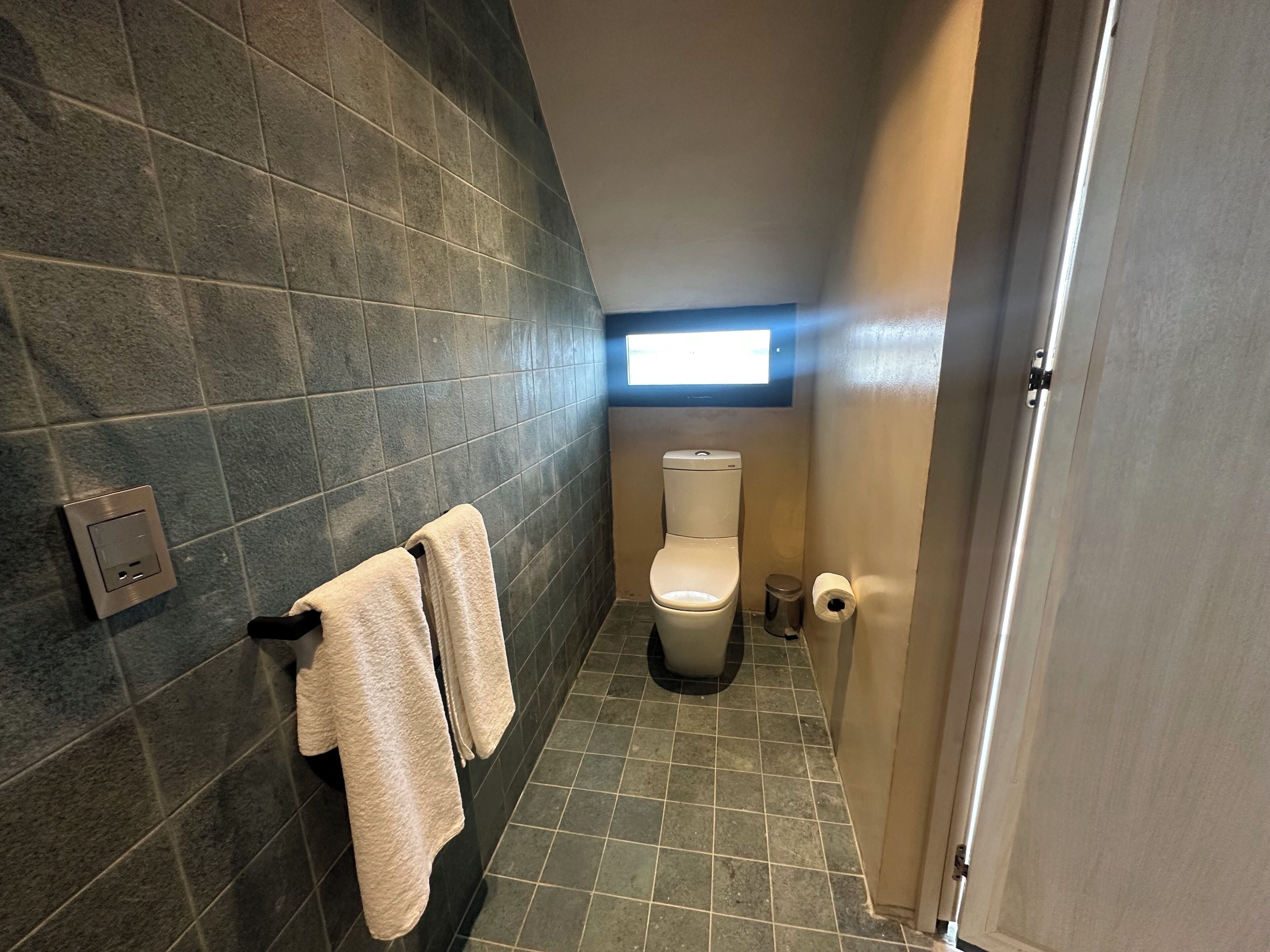 A small bathroom with dark tiles, a toilet, a trash bin, and a towel rack.