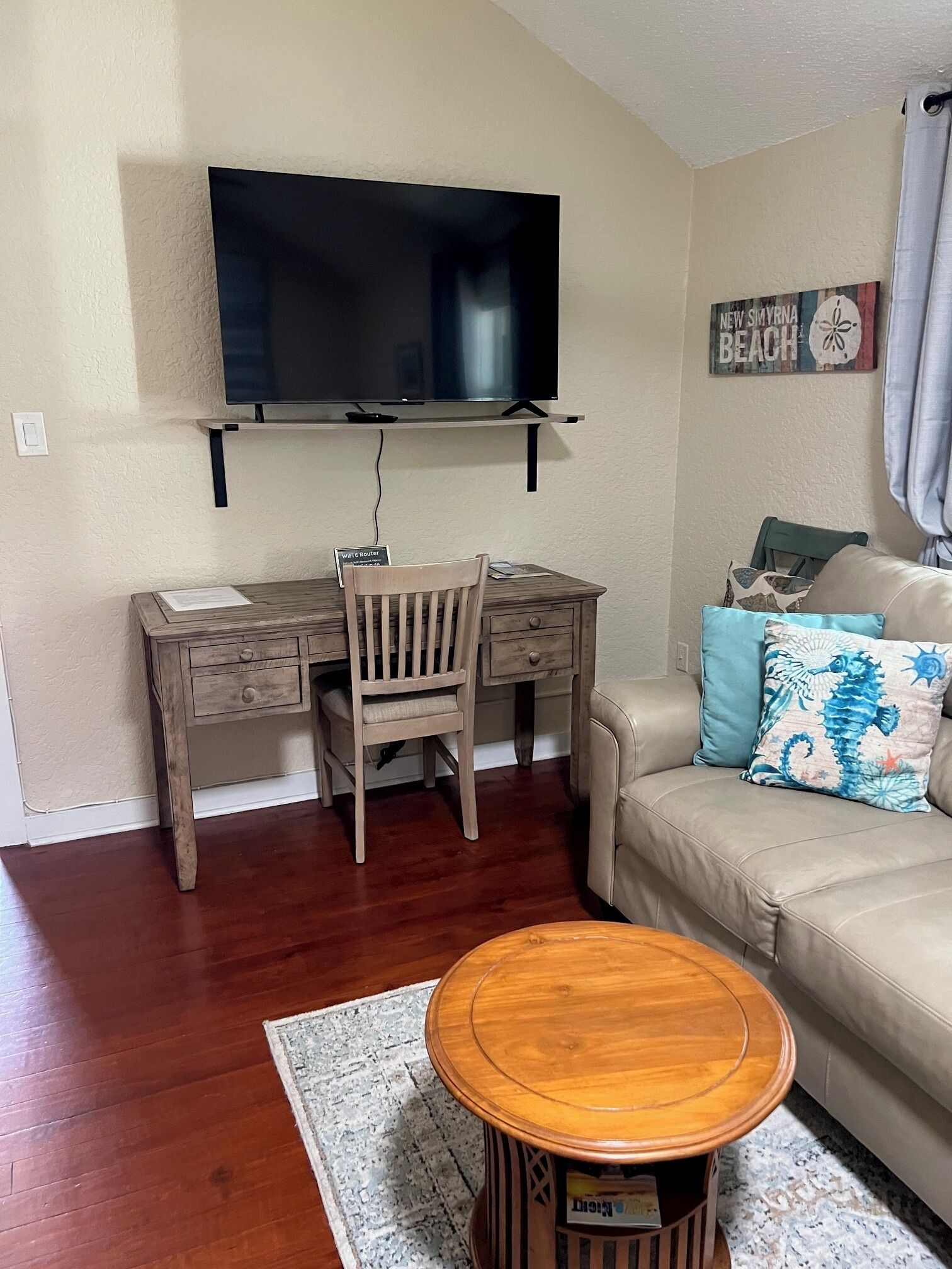 Ein Wohnzimmer mit einem Holztisch, einem Sessel, einem an der Wand befestigten Fernseher, einem Sofa mit Zierkissen und einem runden Couchtisch.