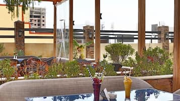 An outdoor seating area with a glass table and chairs, surrounded by plants and a view of buildings.